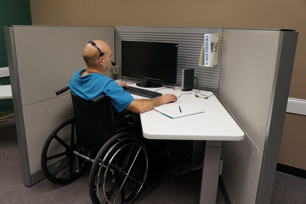 Wheelchair user sitting at a white desk in a call centre with computer screen in front of him and wearing headphones