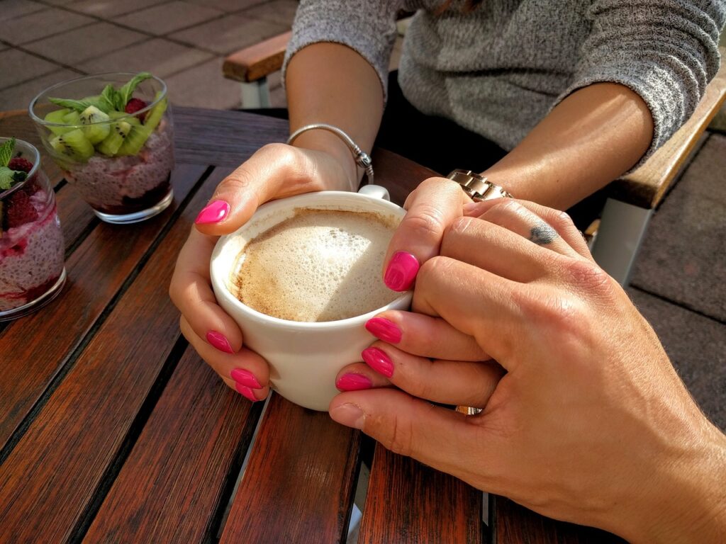 Hands holding a cup of cappuccino with a supportive hand over the holder's left hand. Coffee is resting on a wooden table and only hands of the people photographed are visible. 