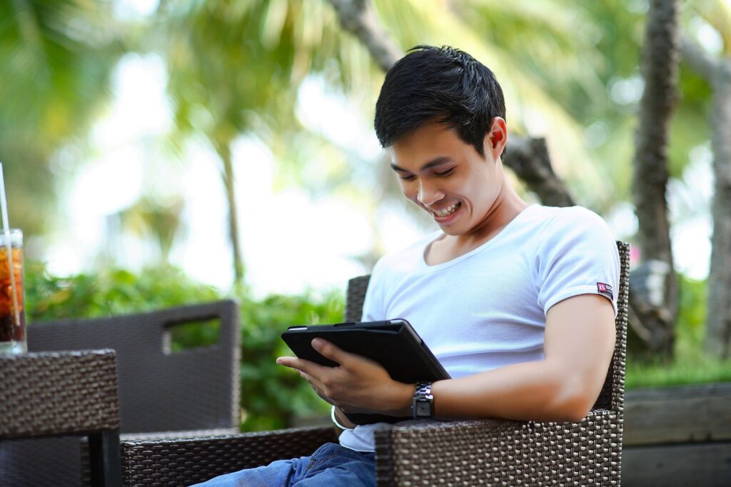Male in a white t-shirt sitting at a table outdoors holding a tablet device and smiling as he interacts with the inclusive business group network for disabled business owners
